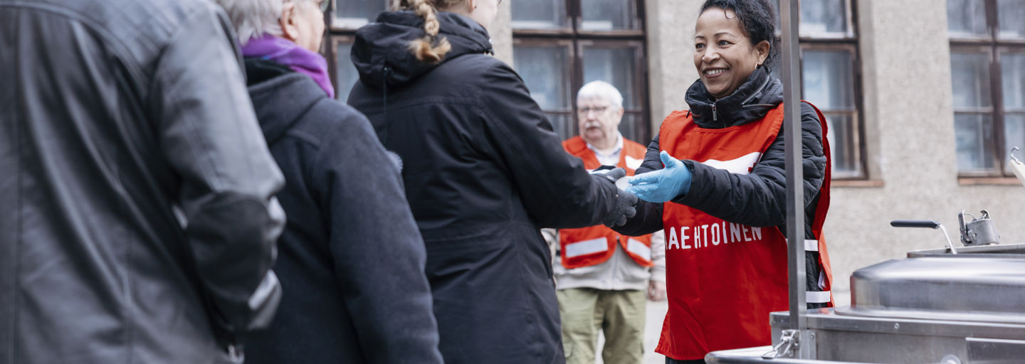 Hymyilevä Punaisen Ristin vapaaehtoinen ojentaa soppa-astiaa ruokajakelujonossa.