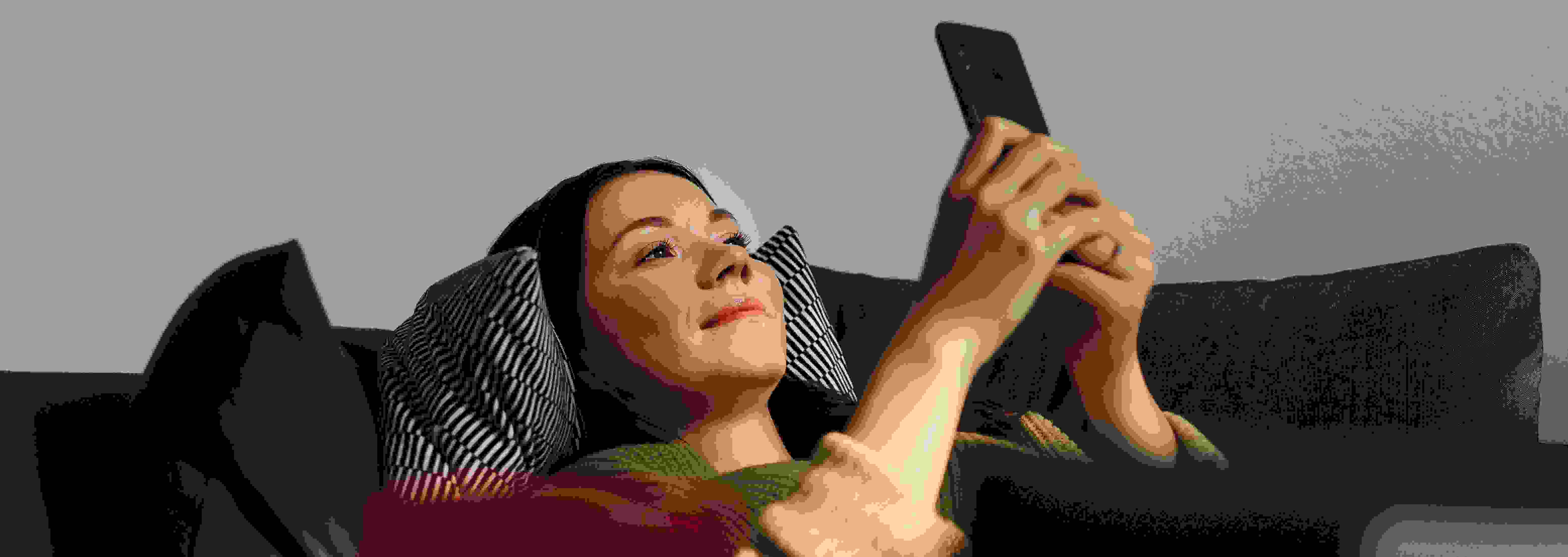 Hymyilevä ihminen makaa selällään sohvalla katsoen älypuhelintaan.