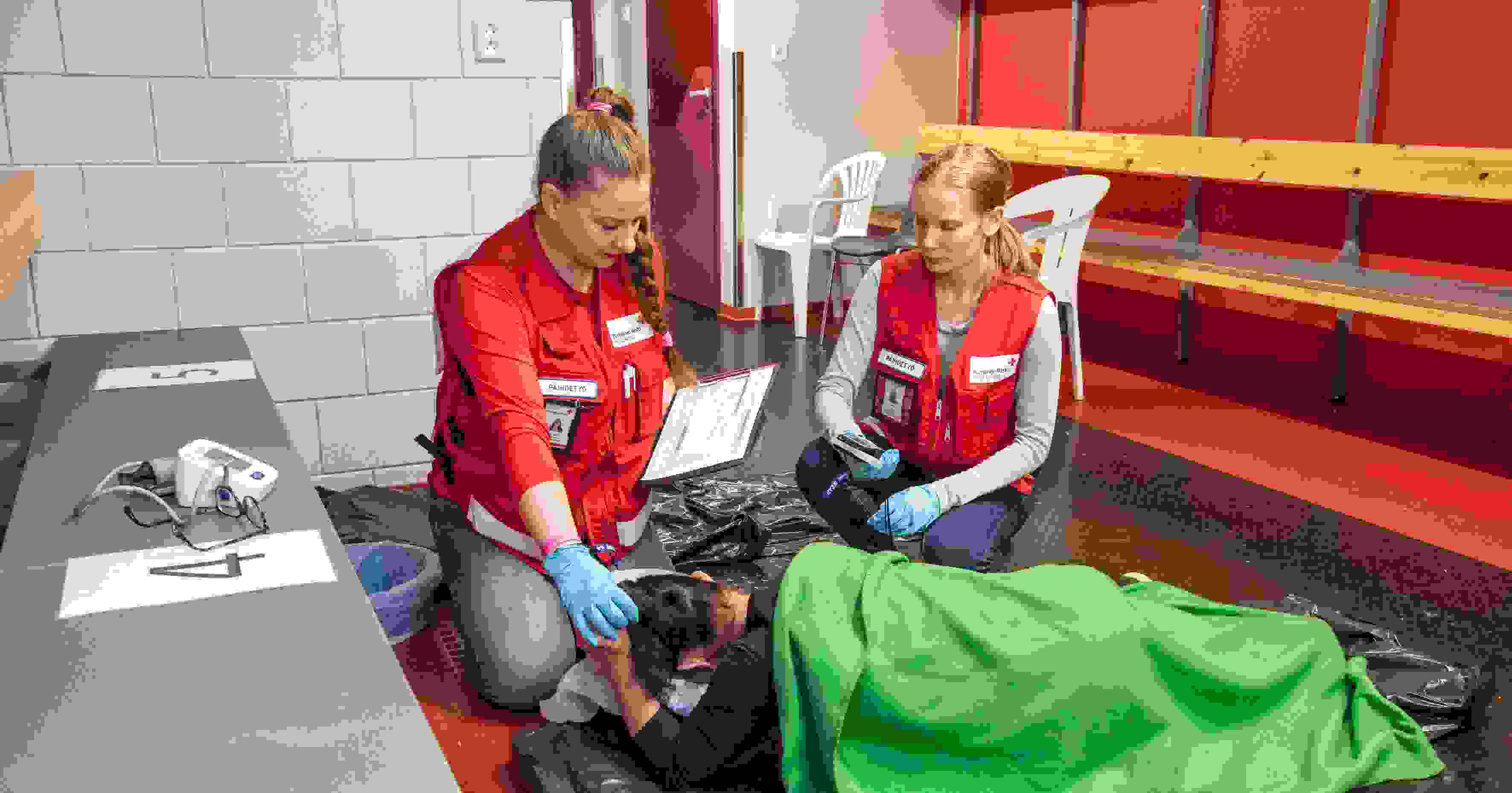 Kuvituskuva Punaisen Ristin tapahtumapäivystyksen selviämispisteestä. Kaksi punaisiin päivystysliiveihin pukeutunutta vapaaehtoista seuraa lattialla lepäävän henkilön vointia.