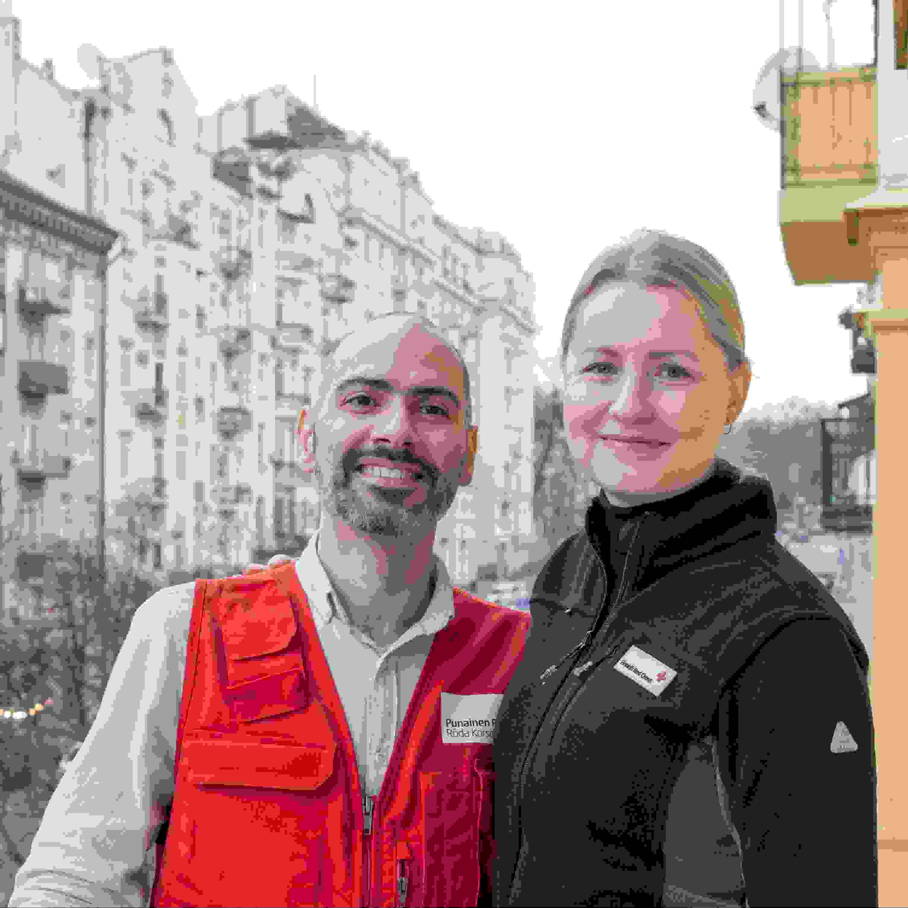 Kaksi hymyilevää Punaisen Ristin avustustyöntekijää kaupunkimaisemassa.