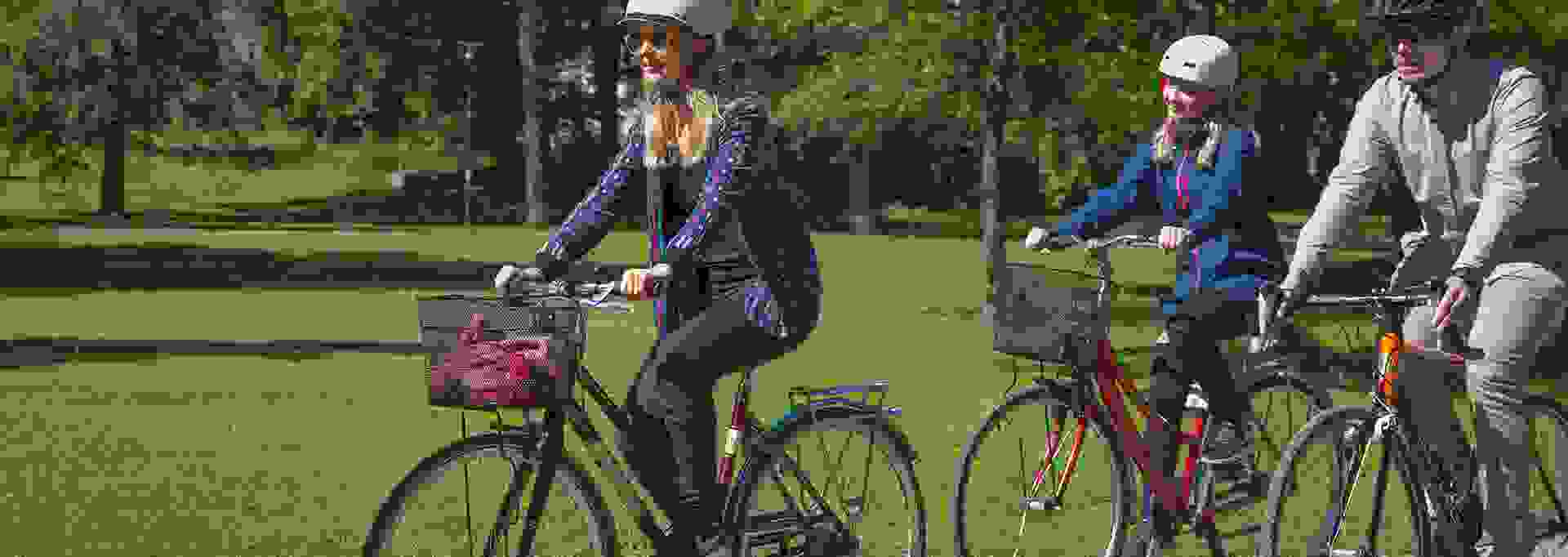 Kolme ihmistä pyöräilee hymyillen puistossa kesäisessä säässä.