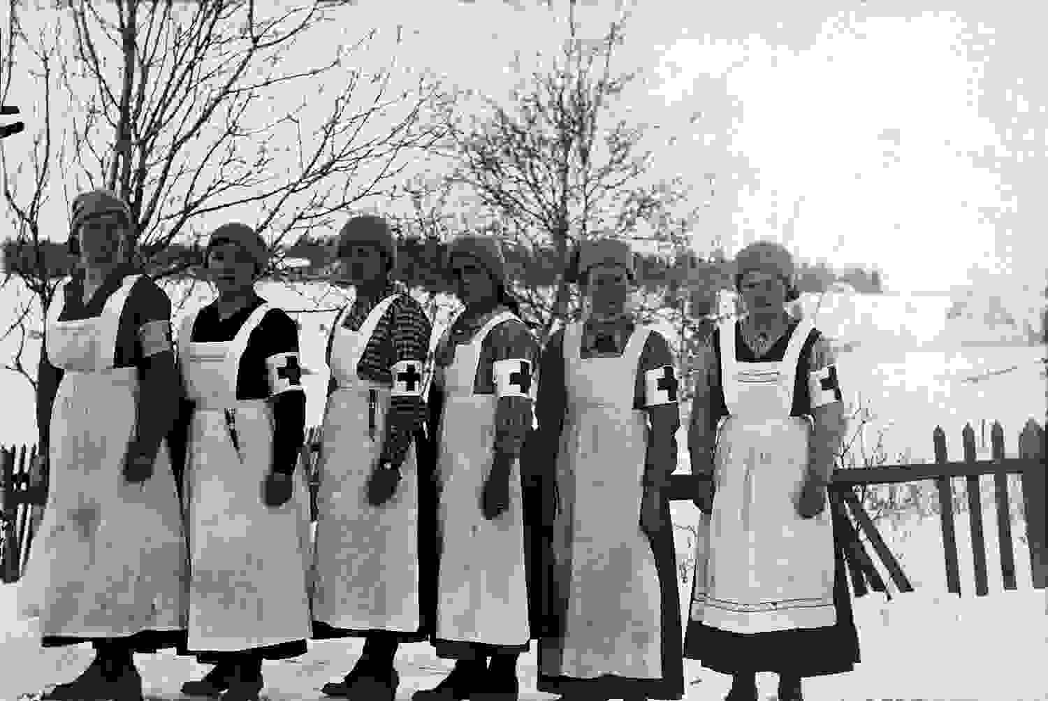 Kuusi vakavailmeistä naista Punaisen Ristin varusteissa seisoo rivissä karussa talvimaisemassa.