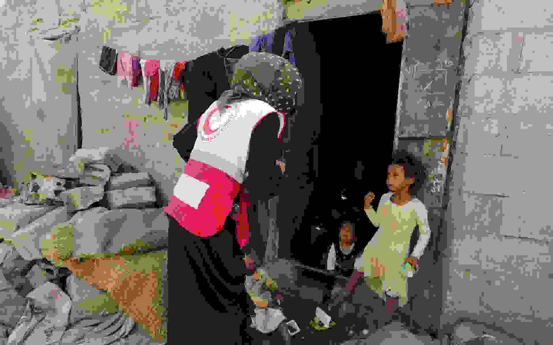 Nainen Punaisen Puolikuun varusteissa juttelee kolmelle pikkulapselle vaatimattoman talon edustalla.