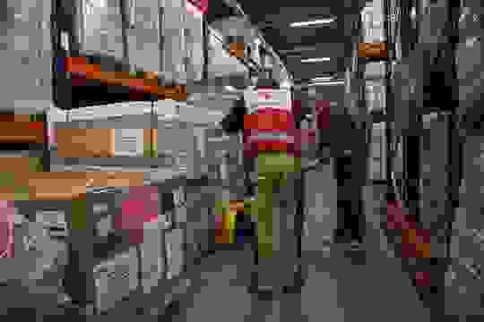 Kaksi Punaisen Ristin työntekijää noutaa avustustarvikkeita logistiikkakeskuksessa, jonka korkeat ja pitkät hyllyt ovat täynnä tarvikkeita.