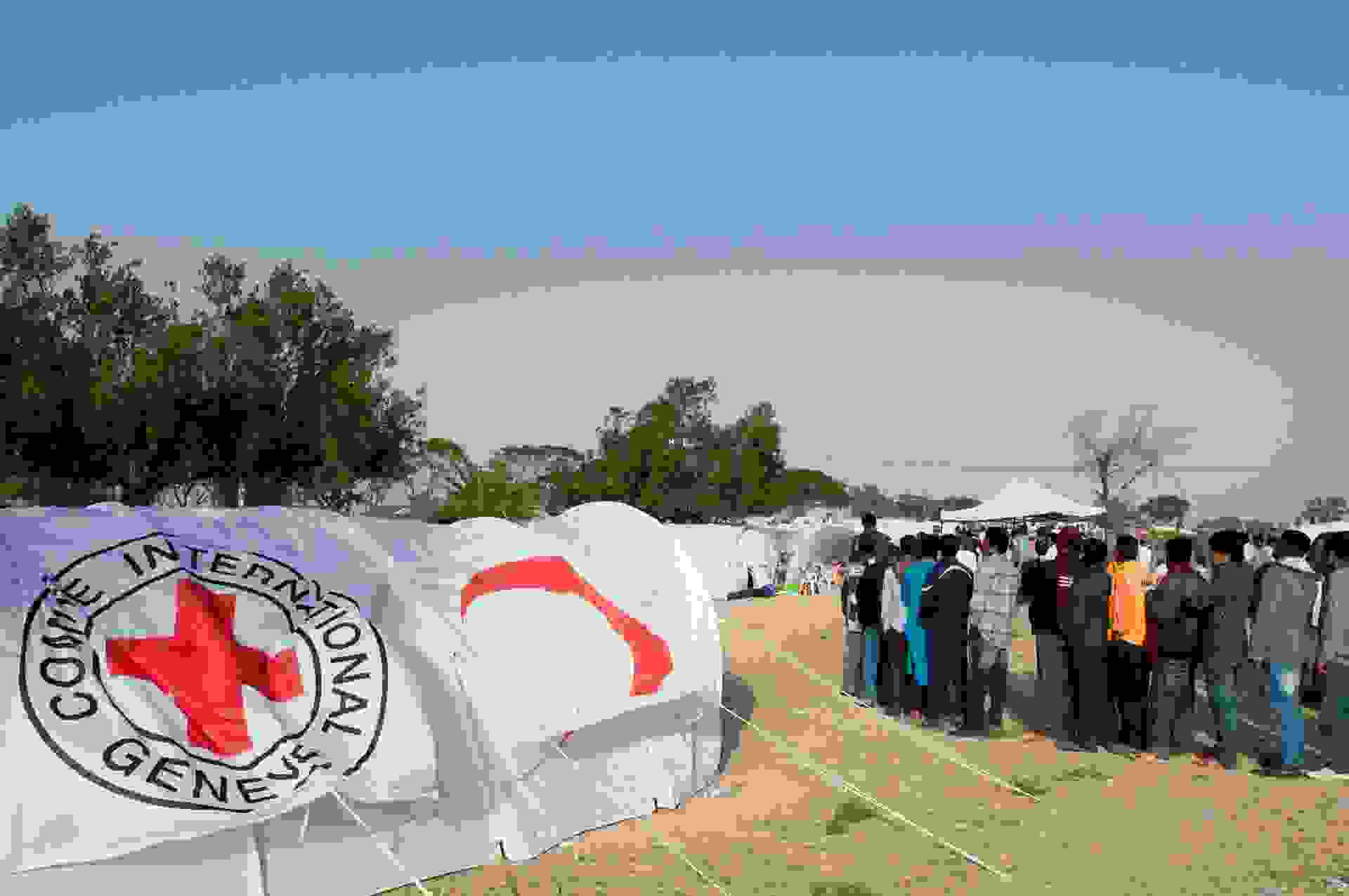 Ihmisiä jonossa pakolaisleirilla Punaisen Ristin tunnuksin varustettujen telttojen keskellä.