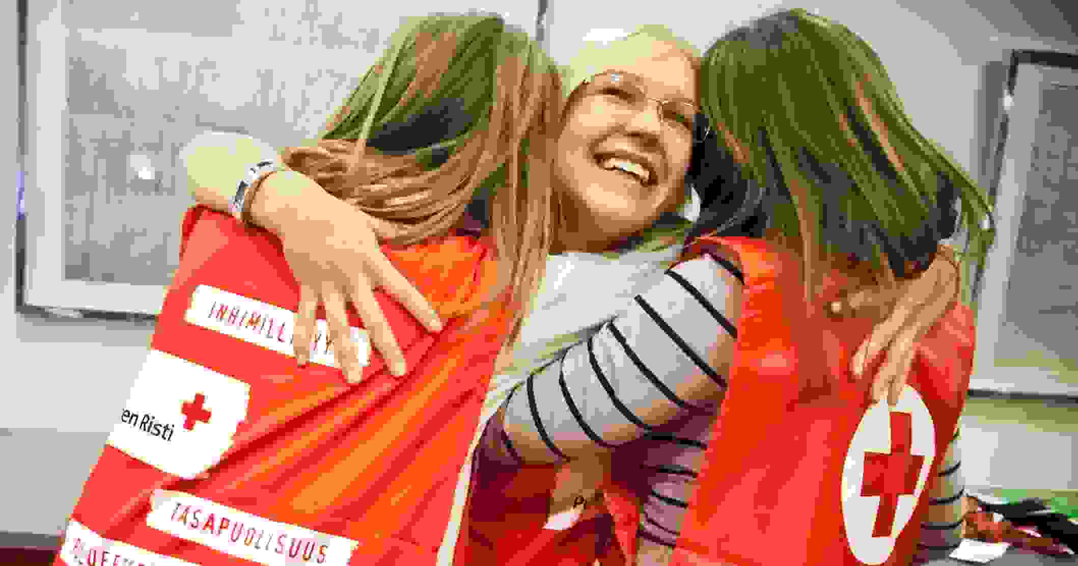 Kolme nuorta naista Punaisen Ristin vapaaehtoisliiveissä hymyilee ja halaa toisiaan.