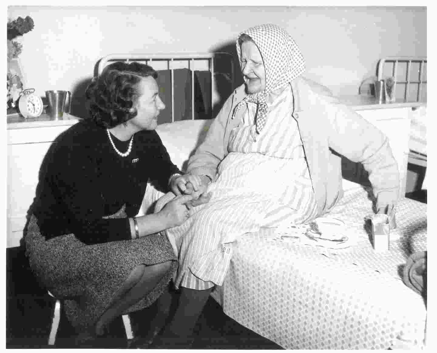 Hymyilevä ikääntynyt nainen ja Punaisen Ristin ystävävapaaehtoinen pitävät eväshetkeä sairaalasängyllä.