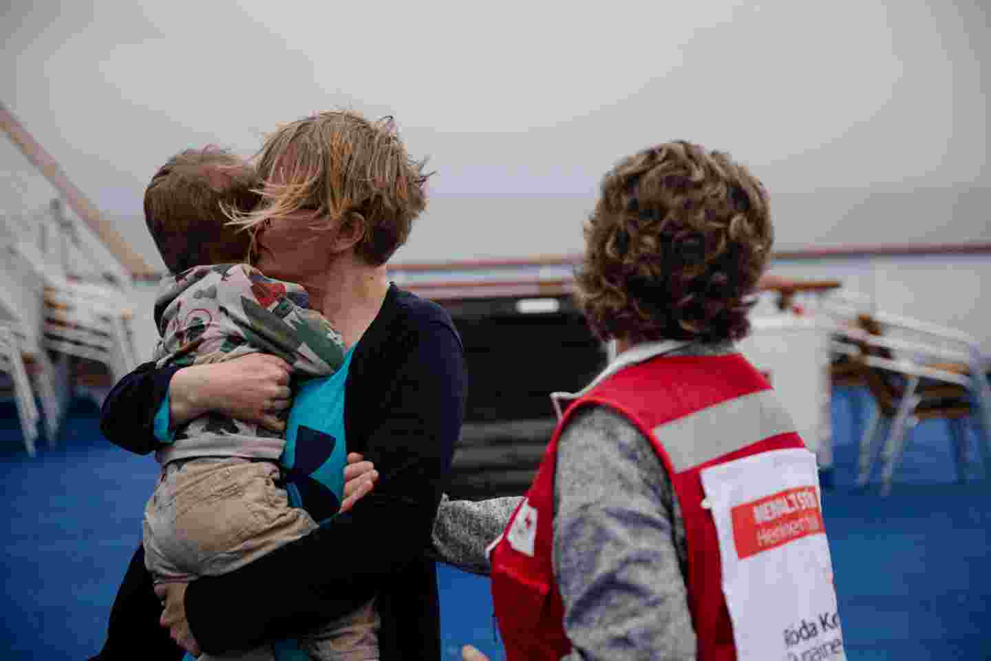 Äiti lapsi sylissään laivan kannella, vieressä Punaisen Ristin vapaaehtoinen.