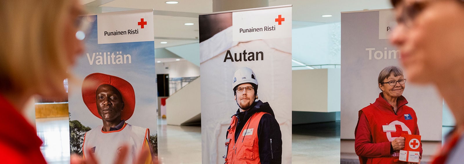 Kaksi henkilöä keskustelee. Taustalla kolme roll-upia, joissa Punaisen Ristin vapaaehtoisia.