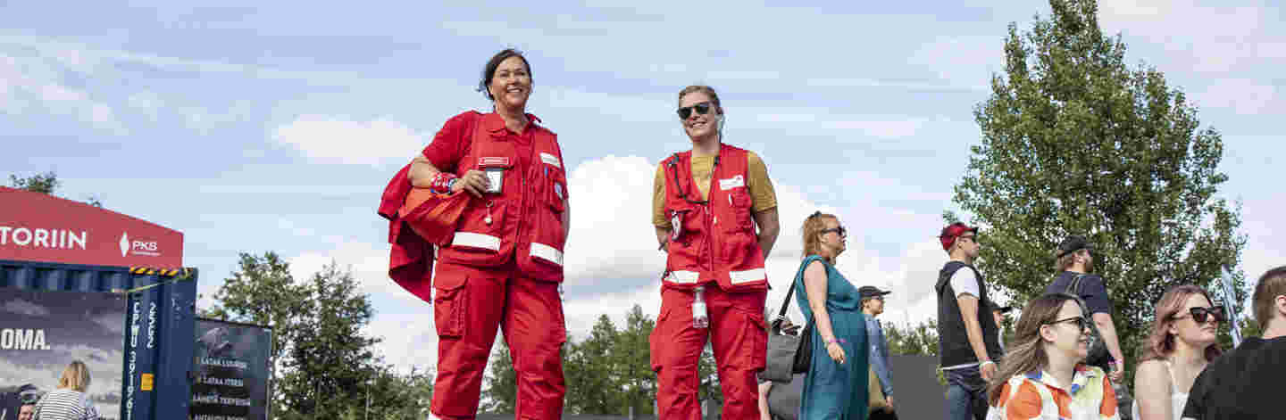Kaksi hymyilevää Punaisen Ristin ensiapupäivystäjää festareilla kesäisessä säässä