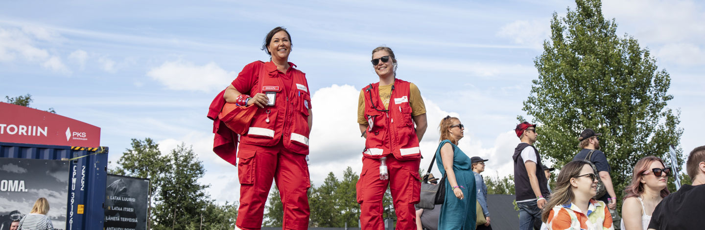Kaksi hymyilevää Punaisen Ristin ensiapupäivystäjää festareilla kesäisessä säässä