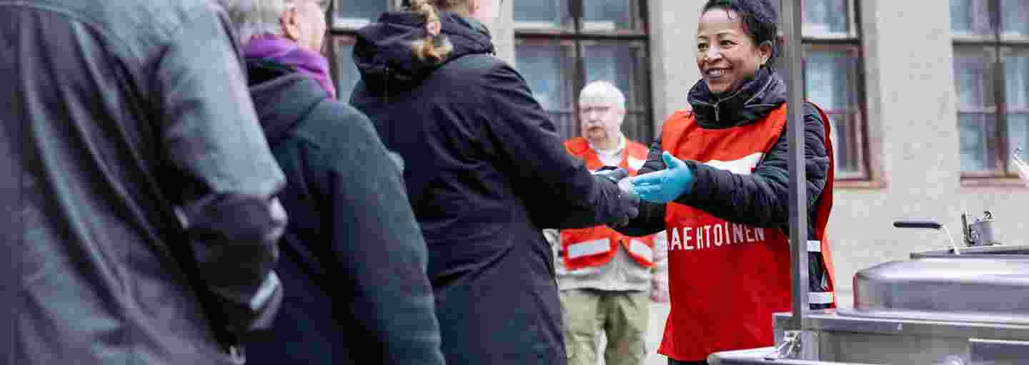 Hymyilevä Punaisen Ristin vapaaehtoinen ojentaa soppa-astiaa avunsaajalle ruokajakelujonossa.