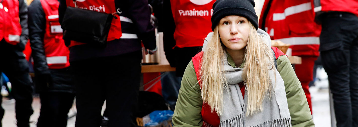 Nuori nainen katsoo eteenpäin yllään Punaisen Ristin vapaaehtoisen liivi. Taustalla on muita vapaaehtoisia.