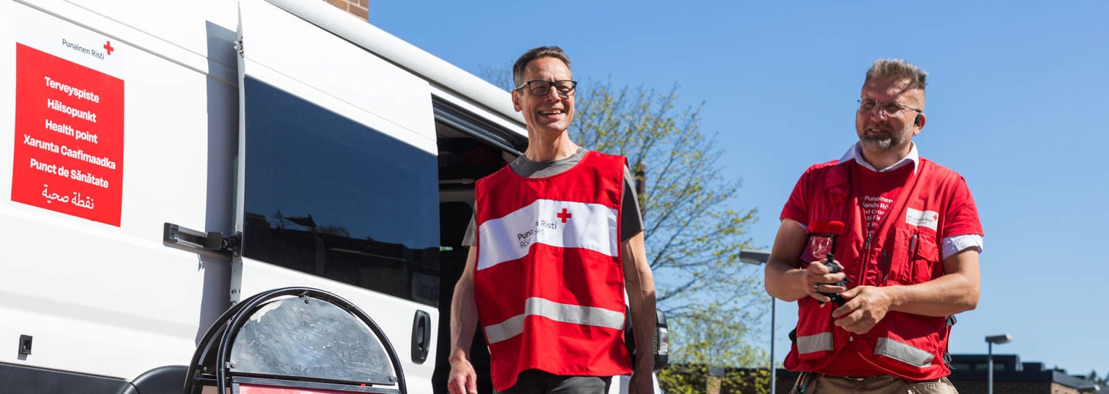 Kaksi hymyilevää Punaisen Ristin vapaaehtoista kävelee keväisessä säässä Punaisen Ristin terveyspisteen pakettiauton vieressä.