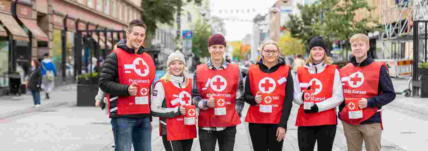 Kuusi nuorta Punaisen Ristin vapaaehtoista lipaskerääjää yllään keräysliivit ja käsissään keräyslippaat. Henkilöt seisovat hymyillen kaupunkimaisemassa.