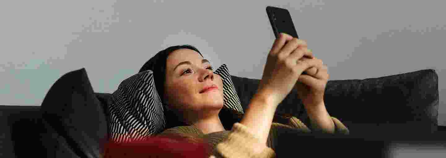Hymyilevä ihminen makaa selällään sohvalla katsoen älypuhelintaan.