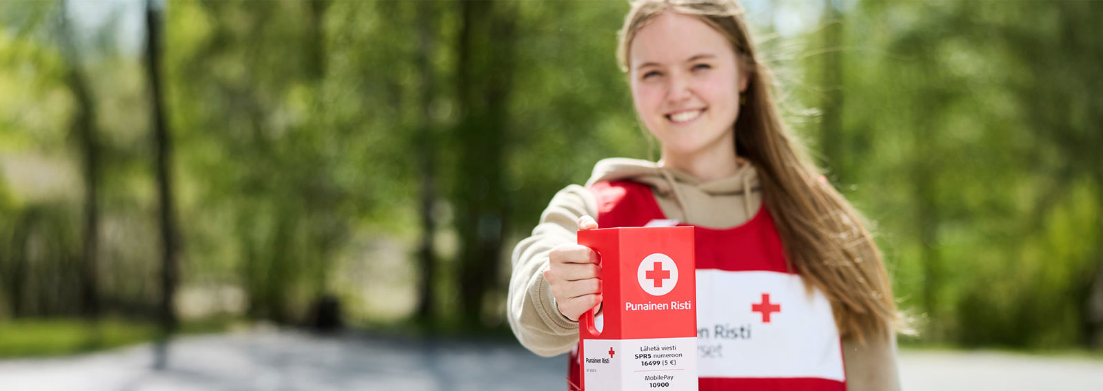 Hymyilevä henkilö yllään Punaisen Ristin keräysliivit ja kädessään keräyslipas