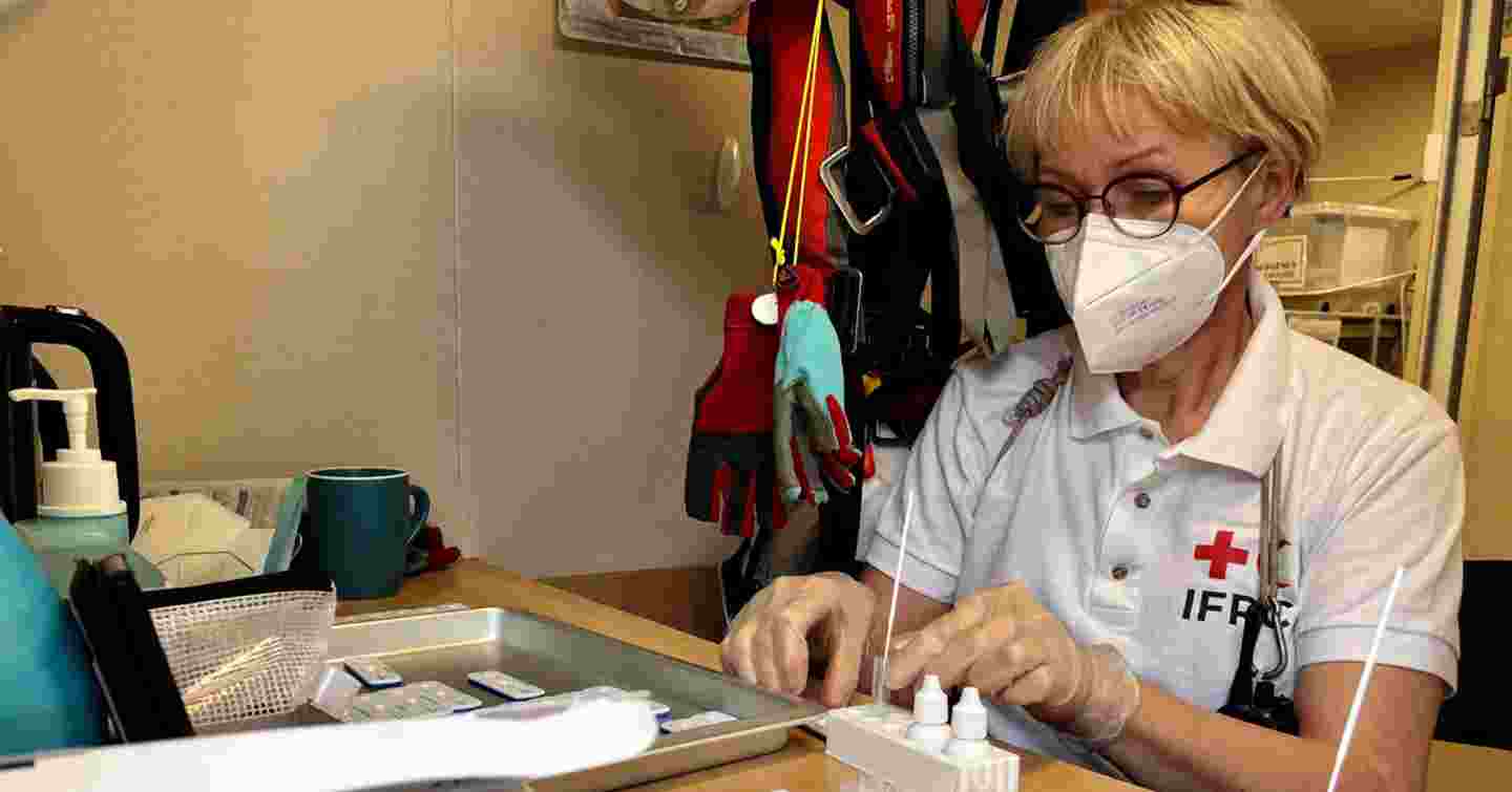 Maskilla suojautunut nainen puolilähikuvassa kirjoituspöydän ääressä käsittelemässä koeputkia.