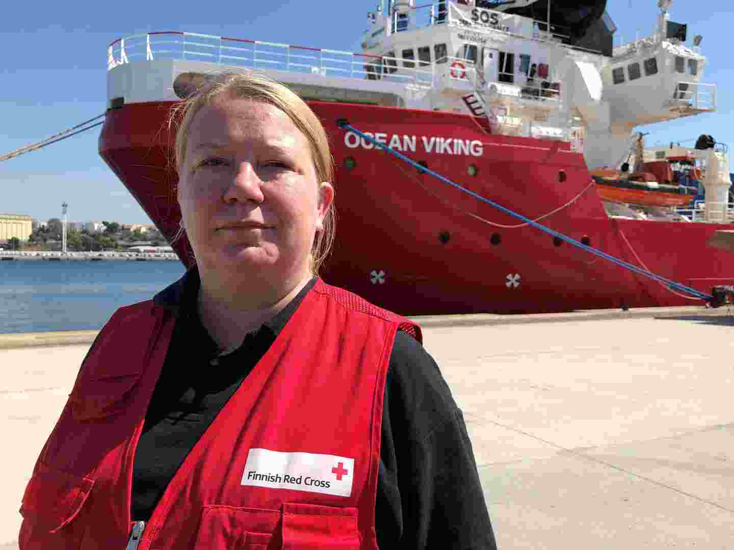 Punaisen Ristin työasuun pukeutunut avustustyöntekijä seisoo laivan edessä satama-alueella.