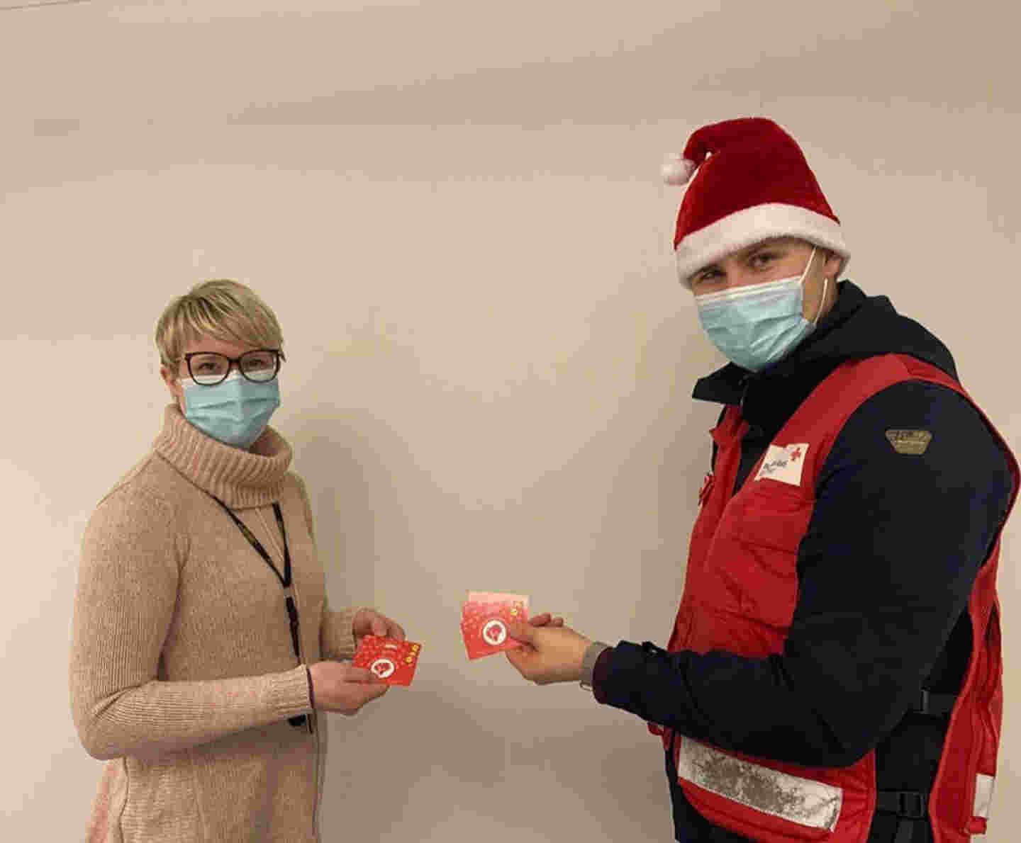 Kaksi henkilöä maskit kasvoillaan pitelee käsissään ruokalahjakortteja. Toisella henkilöistä on yllään Punaisen Ristin liivi ja  päässään tonttulakki.