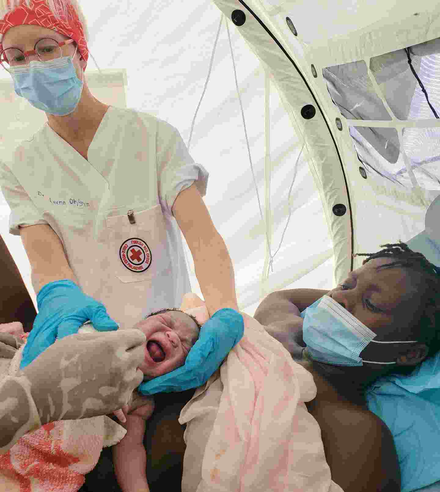 Punaisen Ristin vaatteisiin pukeutunut lääkäri tutkii vastasyntynyttä vauvaa, joka on äitinsä sylissä.