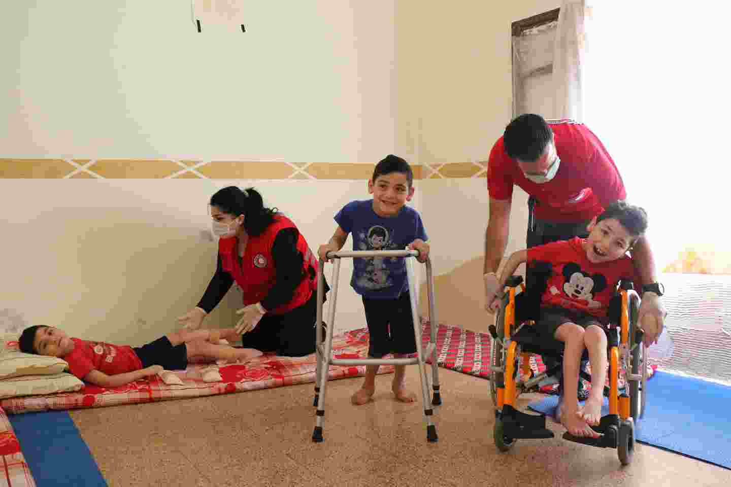 Kolme hymyilevää syyrialaislasta, joita avustaa kaksi Punaisen Puolikuun työntekijää.
