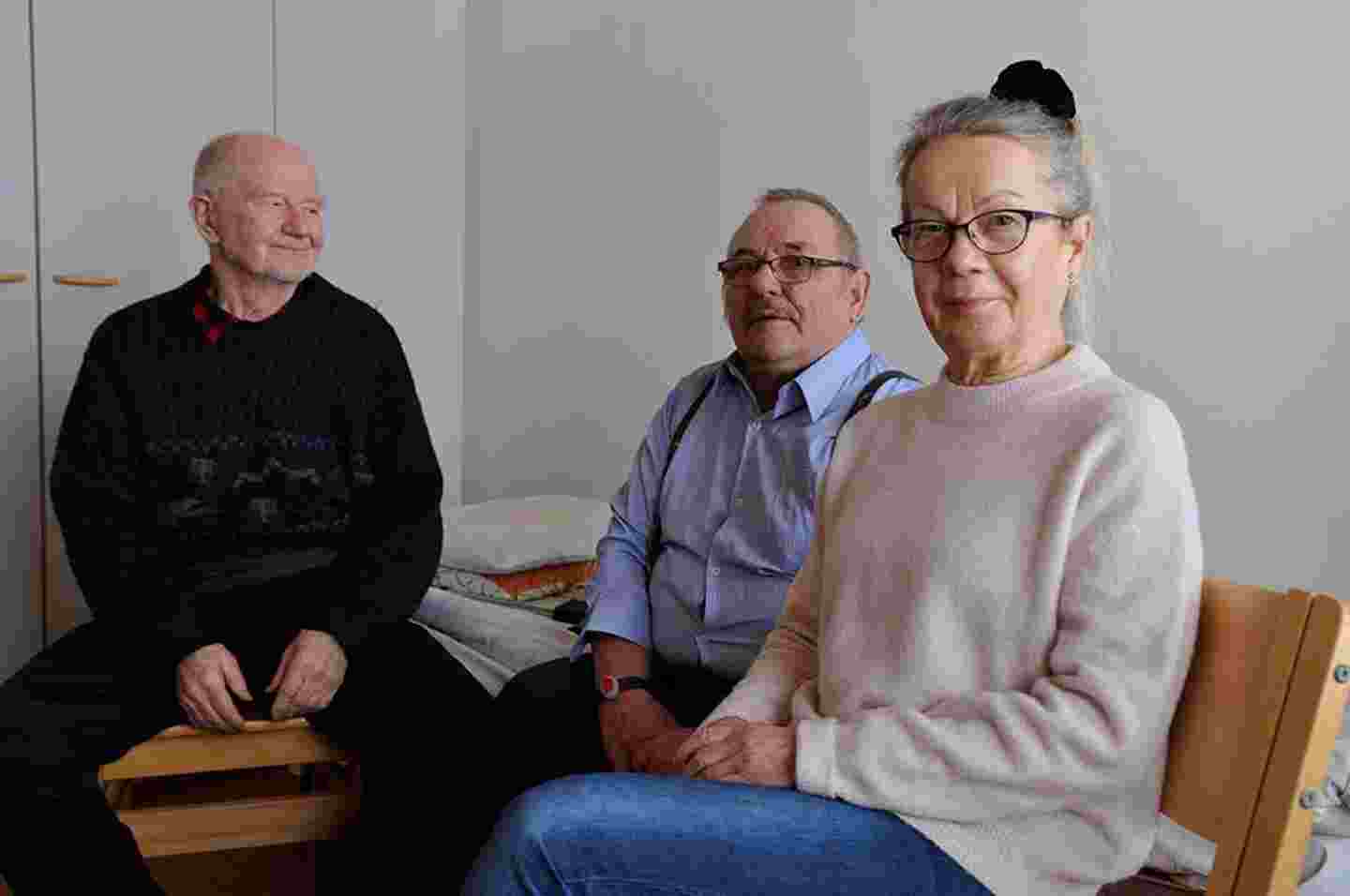 Kolme iäkästä hymyilevää henkilöä istuu vierekkäin.