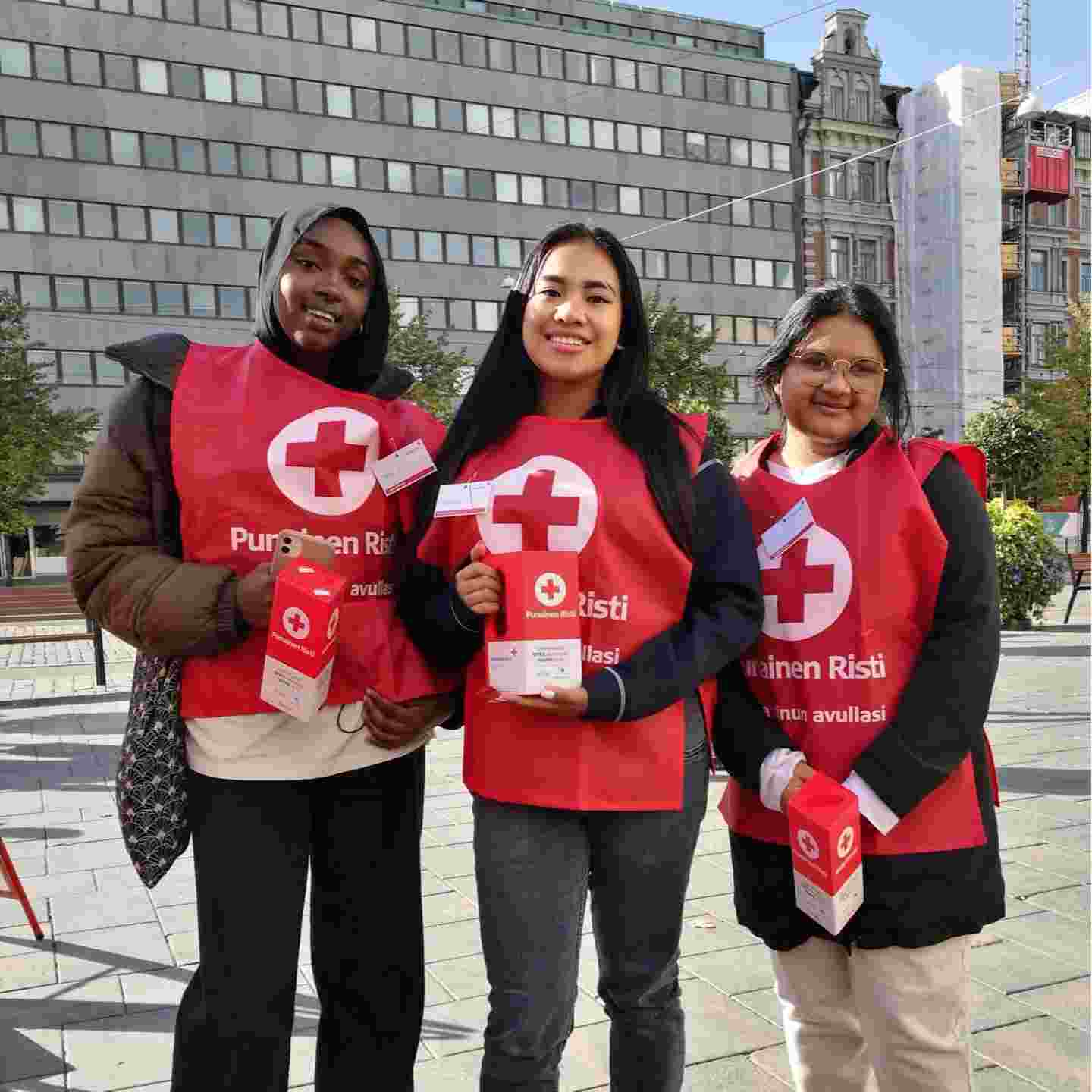 Kolme hymyilevää nuorta yllään Punaisen Ristin liivit ja käsissään keräyslippaat.