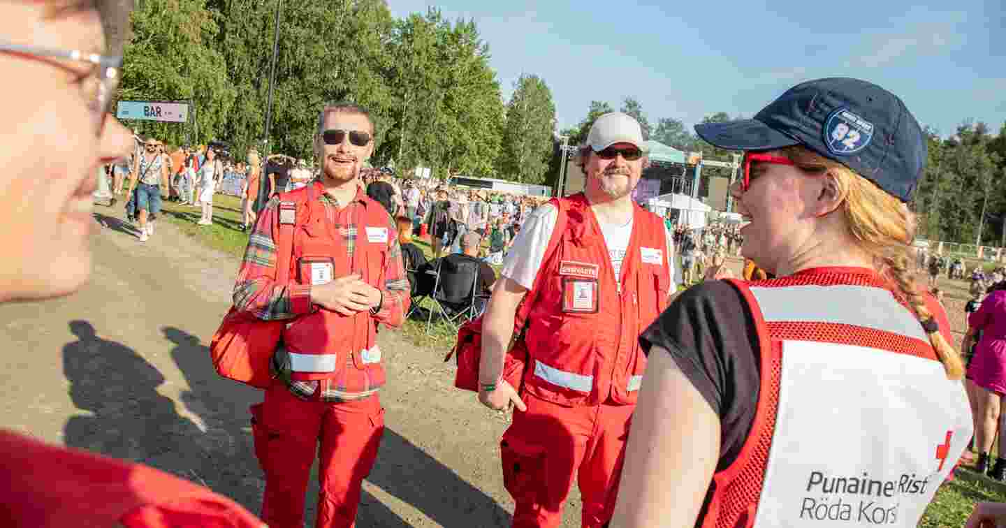 Neljä hymyilevää Punaisen Ristin vapaaehtoista päivystäjää aurinkoisella festivaalialueella.