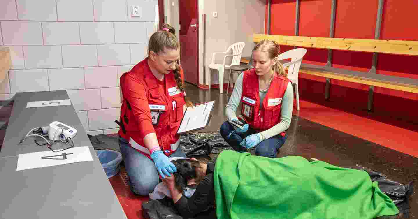 Kuvituskuva Punaisen Ristin tapahtumapäivystyksen selviämispisteestä. Kaksi punaisiin päivystysliiveihin pukeutunutta vapaaehtoista seuraa lattialla lepäävän henkilön vointia.
