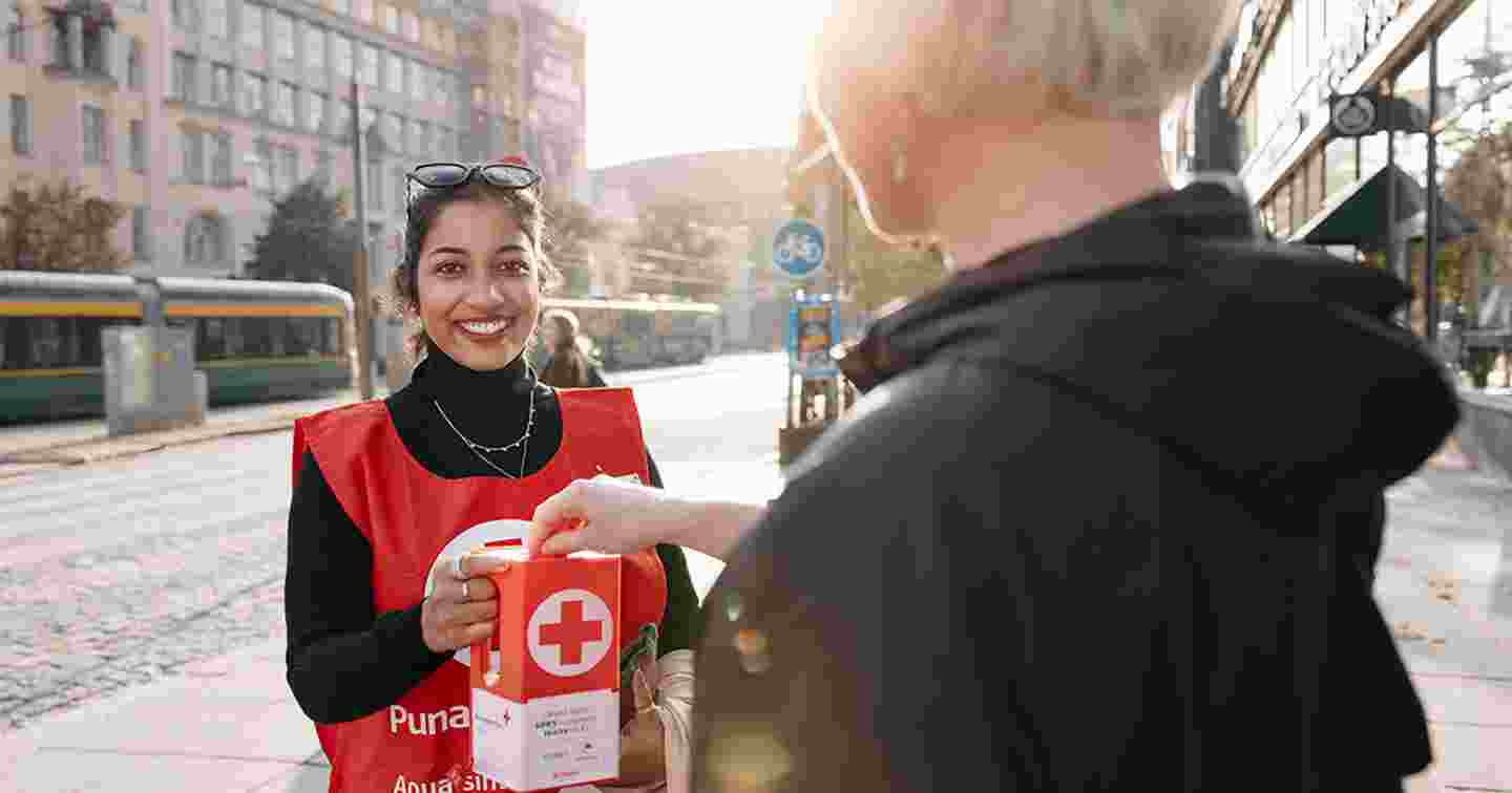 Hymyilevä lipaskerääjä Suomen Punaisen Ristin liiveissä pitelee lipasta, johon ohikulkija laittaa kolikon.
