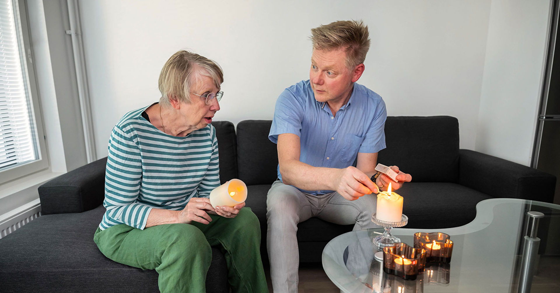 Kaksi ihmistä istuu sohvalla. Toinen sytyttää pöytäkynttilää ja toisella on kädessään led-kynttilä.