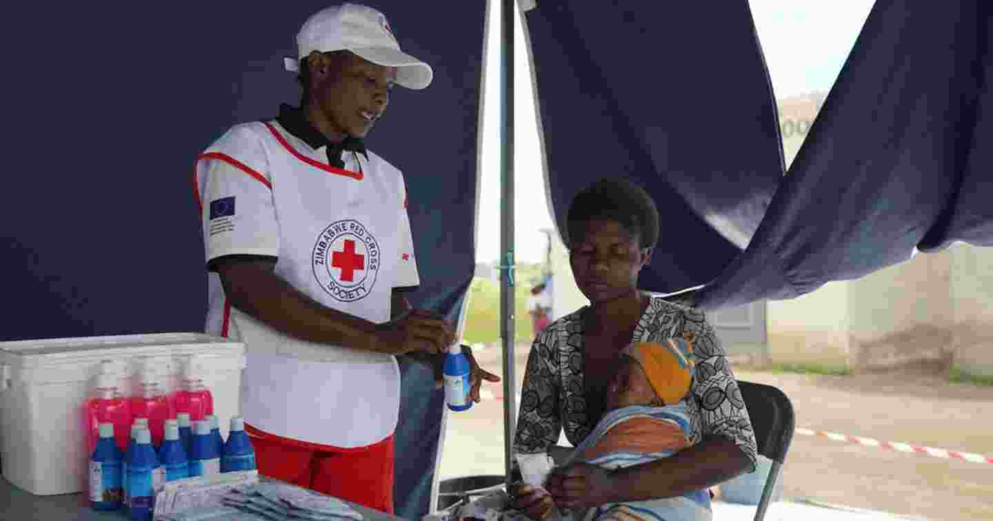 Punaisen Ristin vapaaehtoinen ojentaa vesipulloa tuolilla istuvalle henkilölle ja lapselle.