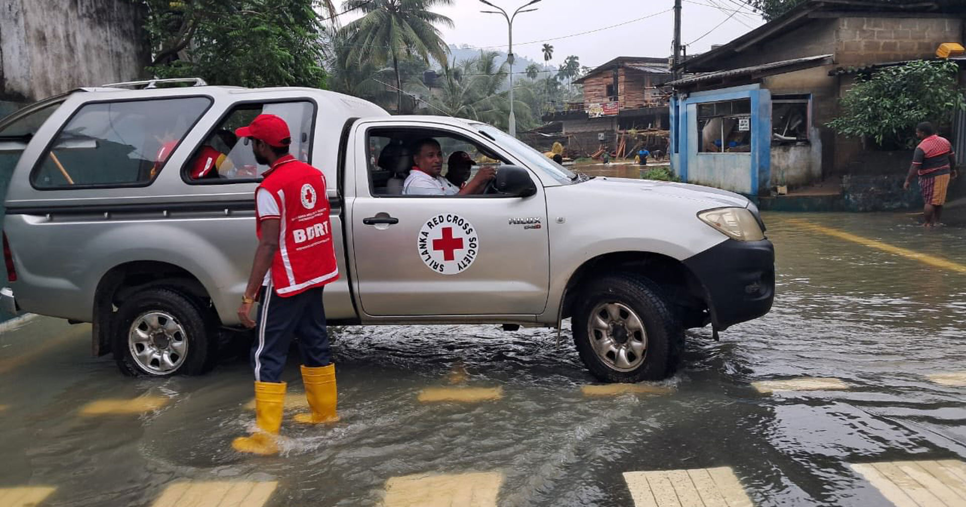 Punaisen Ristin avustustyöntekijä seisoo tulvavedessä keltaisissa saappaissa ja punaisessa liivissä, vierellään harmaa avustusajoneuvo, jossa on Punaisen Ristin logo ovessa. Auto on osittain veden peittämällä kadulla, ja taustalla näkyy trooppisia puita, sähkölinjoja sekä rakennuksia tulva-alueella.