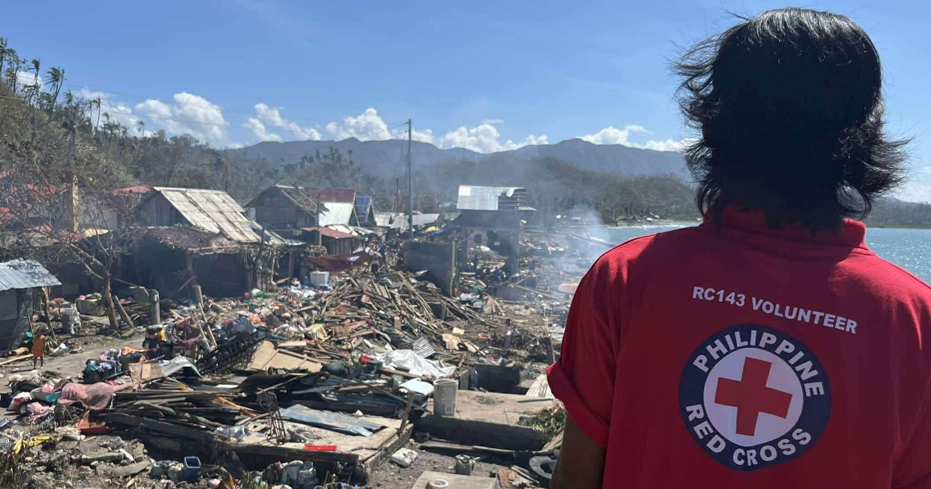 Filippiinien Punaisen Ristin vapaaehtoinen katsoo rantamaisemaa, joka on täynnä myrskytuhoja, kuten tuhoutuneita taloja.