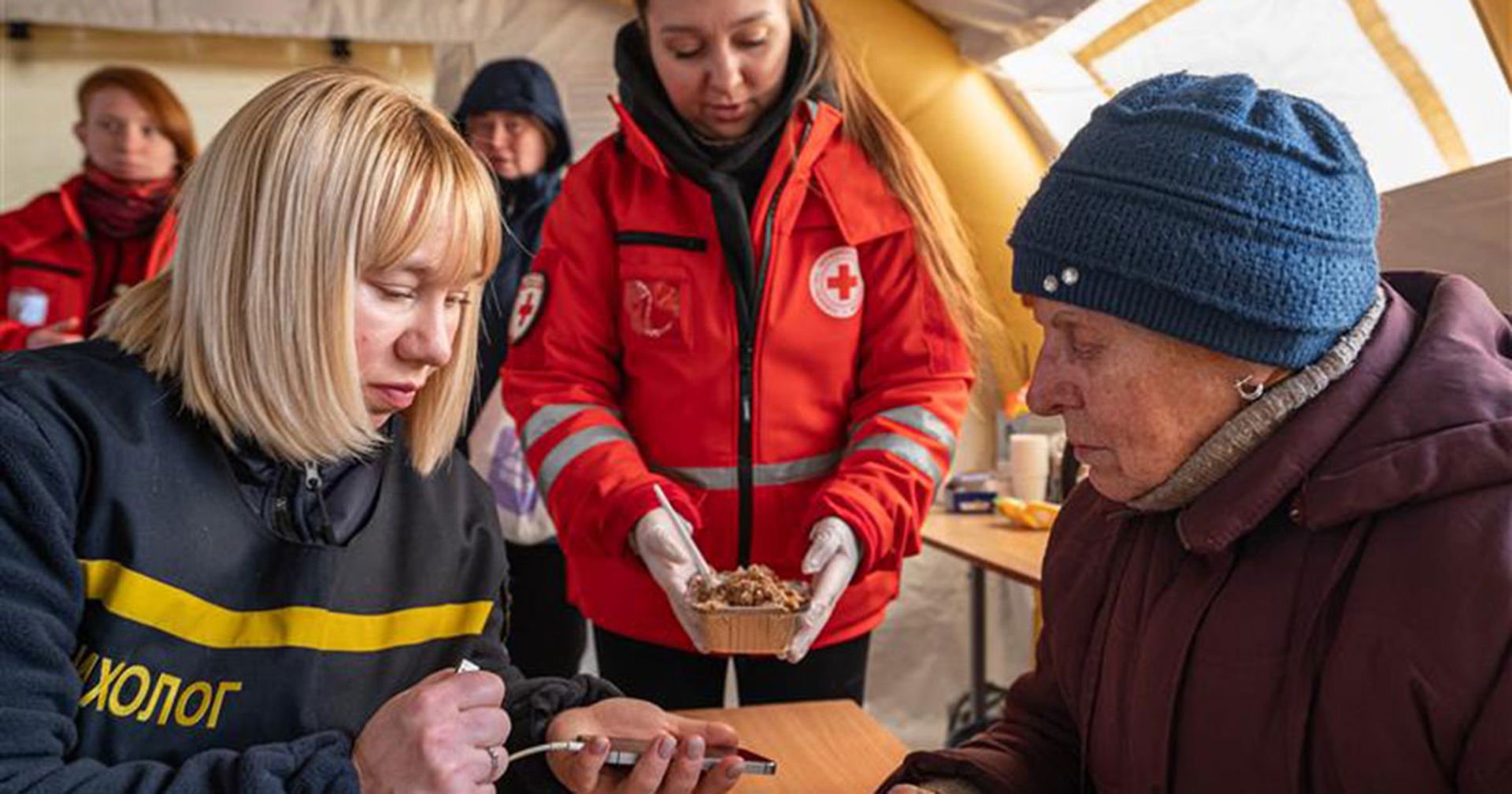 Ukrainan Punaisen Ristin lämmityspiste Kiovassa. Kaksi henkilöä istuu pöydän ääressä ja heistä toinen lataa puhelinta. Ukrainan Punaisen Ristin vapaaehtoinen seisoo taustalla ja tarjoaa ruokaa.