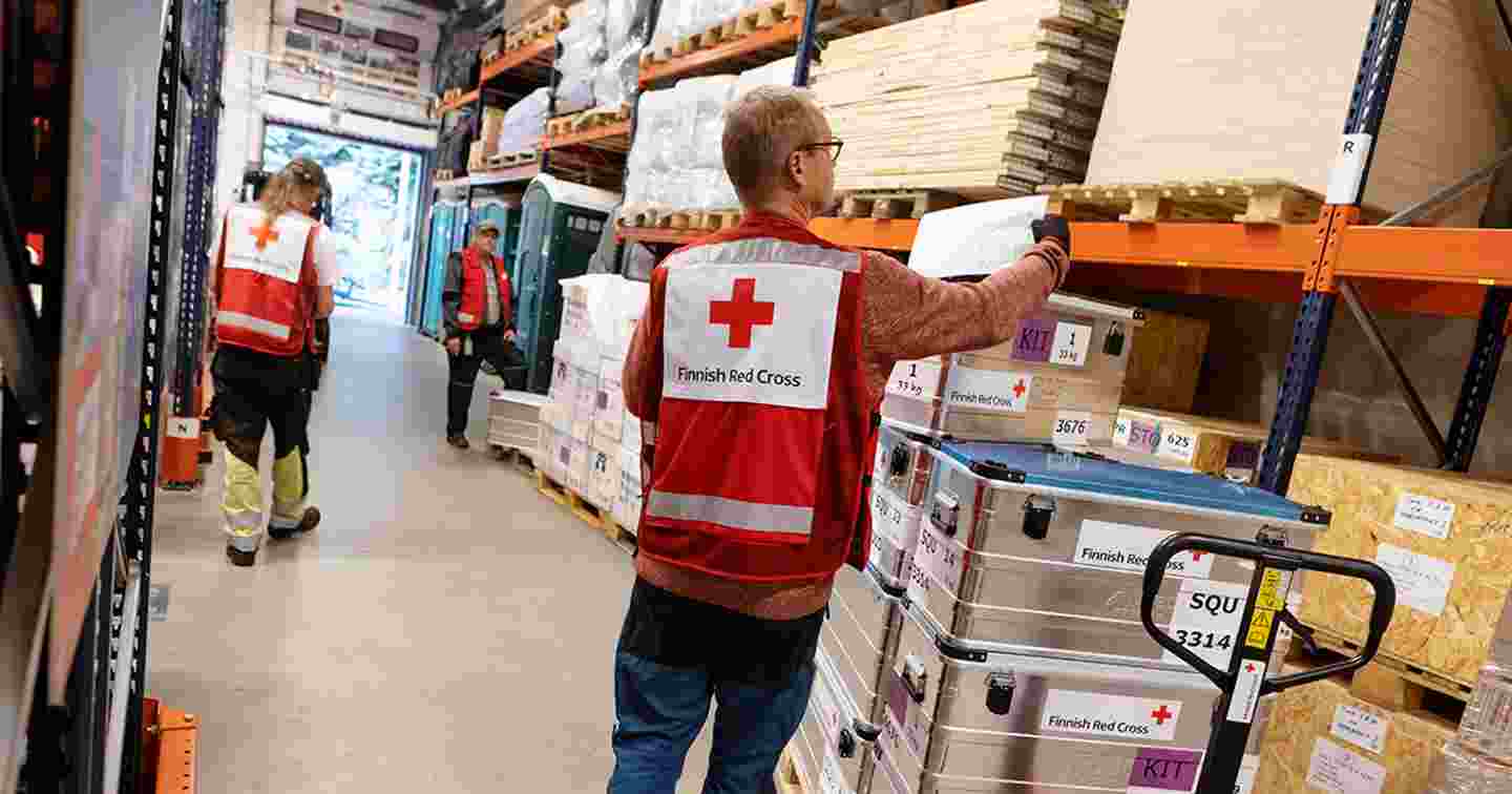 Kolme miestä Punaisen Ristin vaatteissa pakkaavat kenttäsairaalavarusteita isossa varastorakennuksessa.