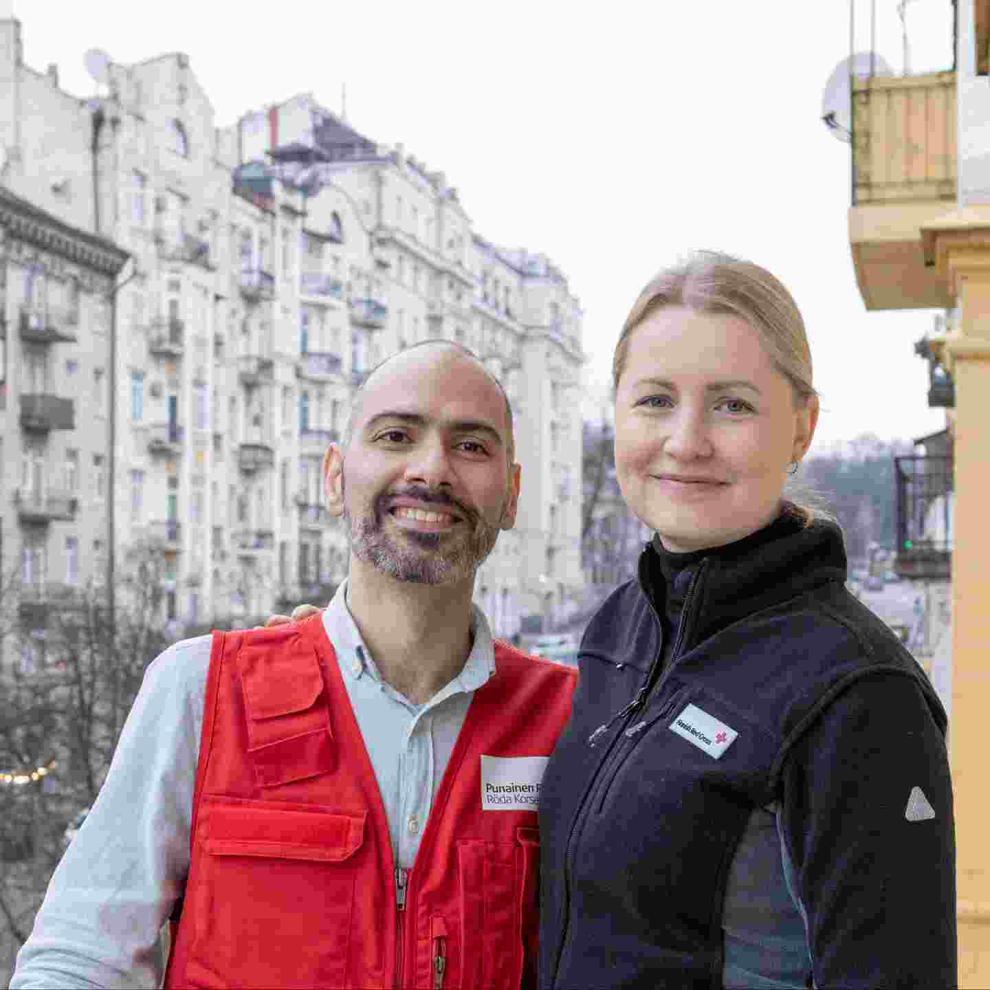 Kaksi hymyilevää Punaisen Ristin avustustyöntekijää kaupunkimaisemassa.