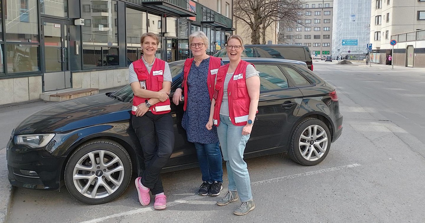 Kolme hymyilevää henkilöä Punaisen Ristin vaatteissa seisoo auton edessä.