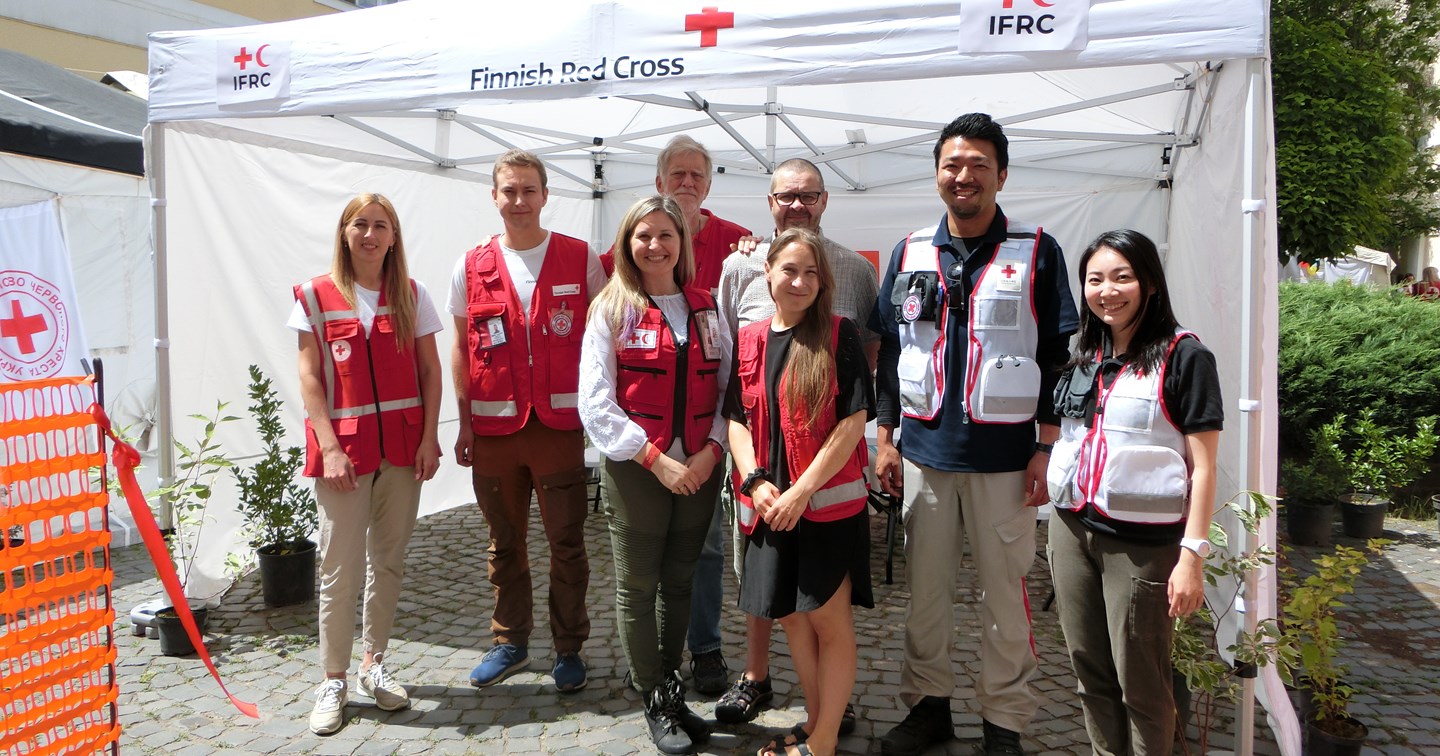 Kahdeksan avustustyöntekijää Punaisen Ristin työvaatteissa seisoo hymyillen terveysasemaan kuuluvan teltan edessä.