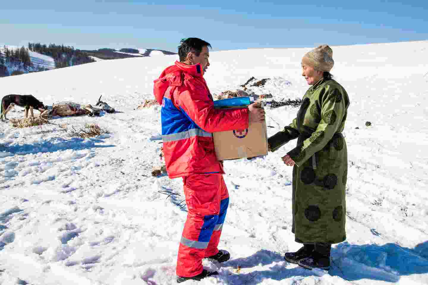 Mies Punaisen Ristin varusteissa ojentaa avustustarvikkeita miehelle lumisessa vuoristomaisemassa.
