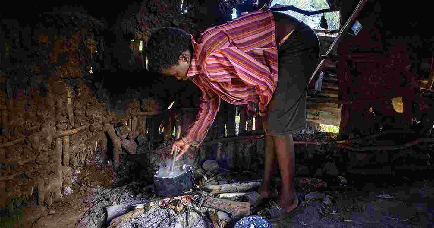 Nuori nainen valmistaa ruokaa nuotiolla maapohjaisessa majassa, jonka seinässä on reikiä.