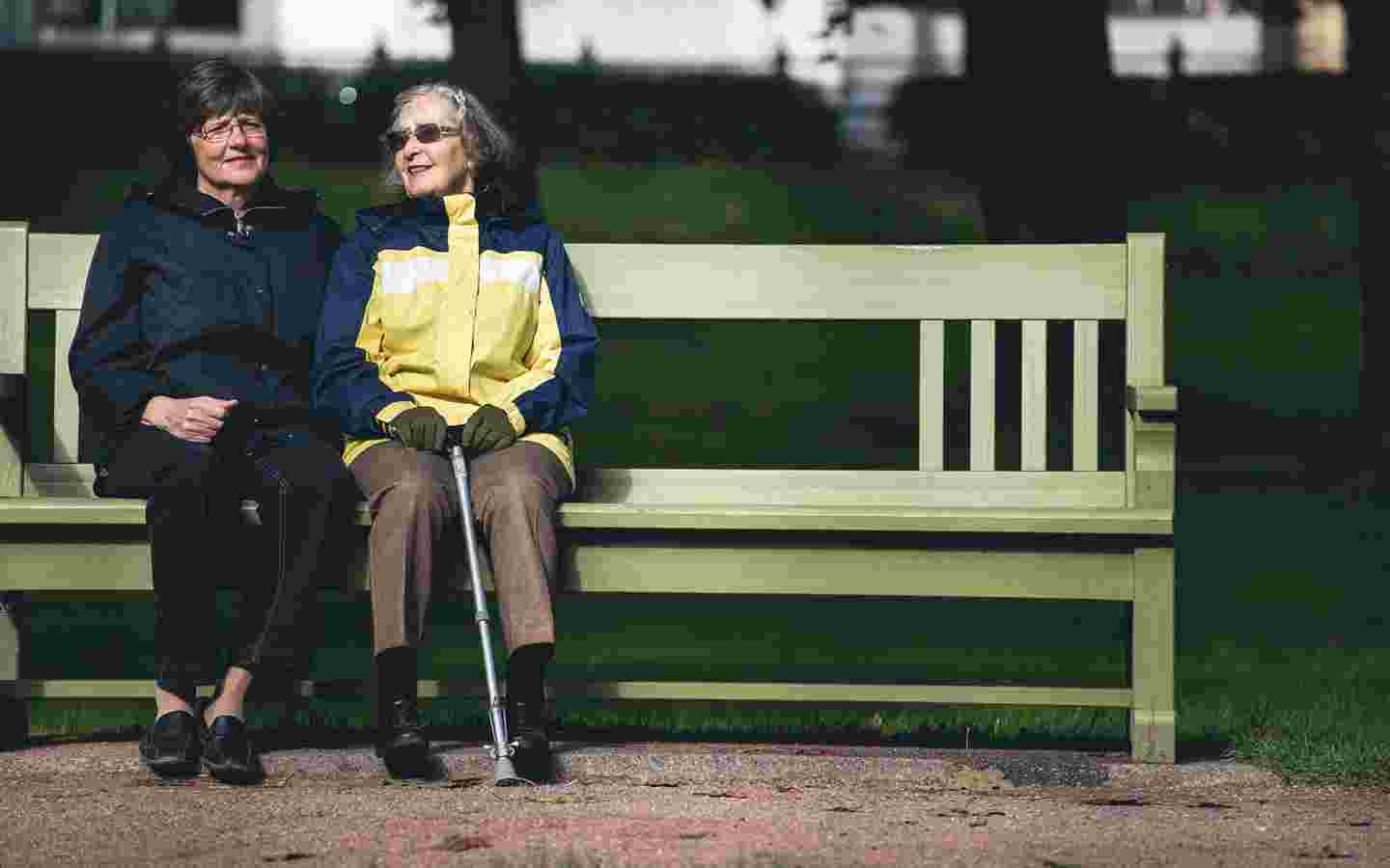 Kaksi ikääntynyttä naista istuu vierekkäin puistonpenkillä.