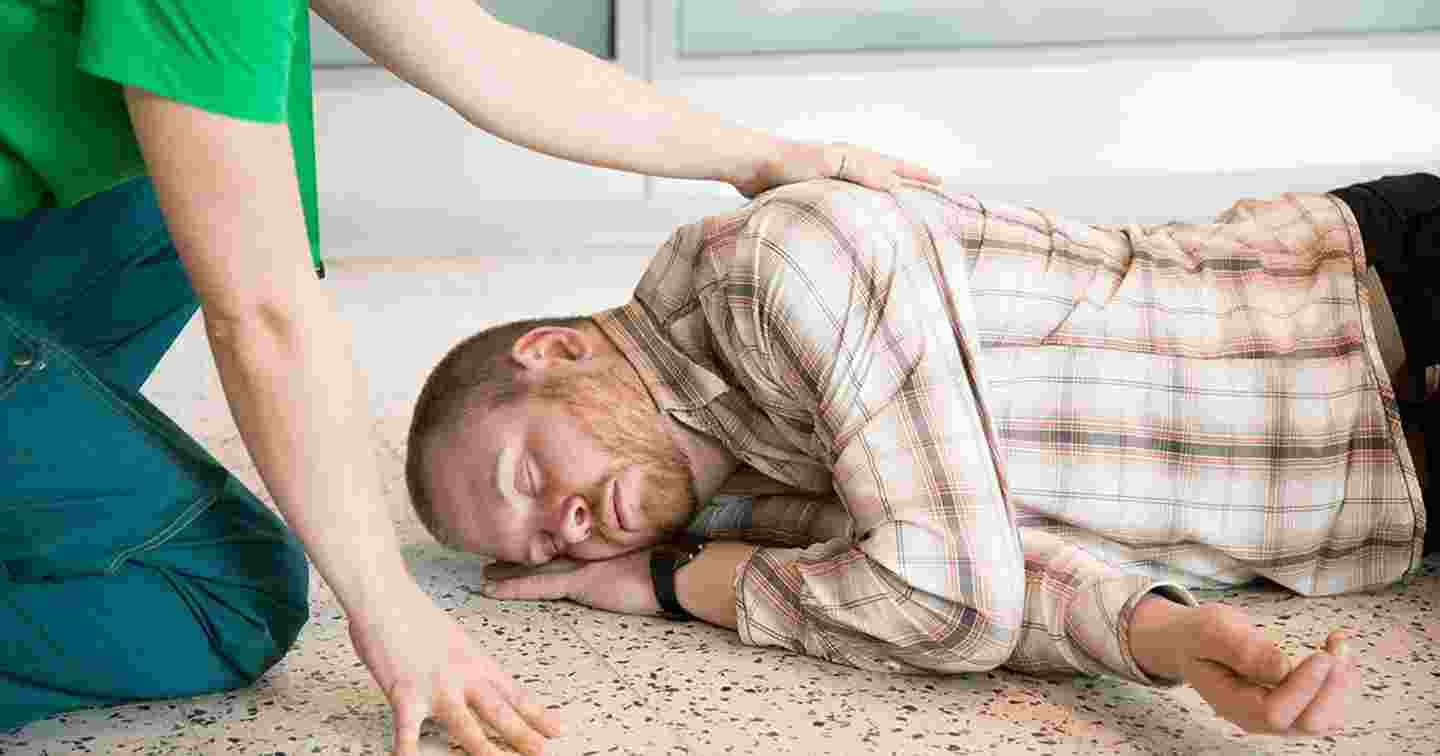 Henkilö makaa silmät suljettuina kylkiasennossa lattialla vieressään auttaja, joka pitää kättä tämän olkapäällä.