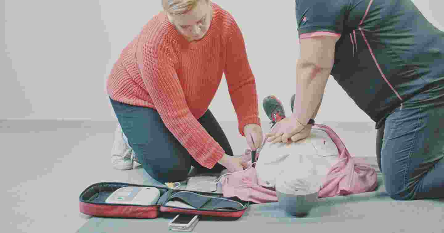 Kaksi henkilöä polvillaan lattialla harjoittelee defibrillaattorilla elvytystä elvytysnuken avulla.