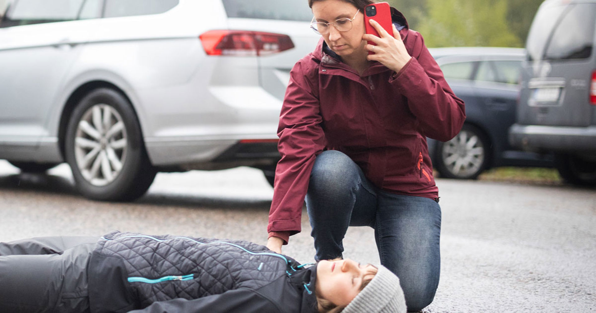 Henkilö on polvistunut tajuttoman lapsen viereen ulkona alfaltilla. Hän pitää toisella kädellään lasta olkapäästä ja soittaa toisella kädellään hätänumeroon puhelimella. Taustalla näkyy pysäköityjä autoja.  lasta olkapäästä ja soittaa toisella 
