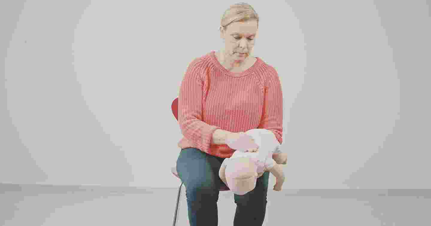 Henkilö istuu tuolilla pitäen käsissään vauva-elvytysnukkea ja iskee tätä selkään harjoitellen vierasesineen poistamista hengitysteistä.