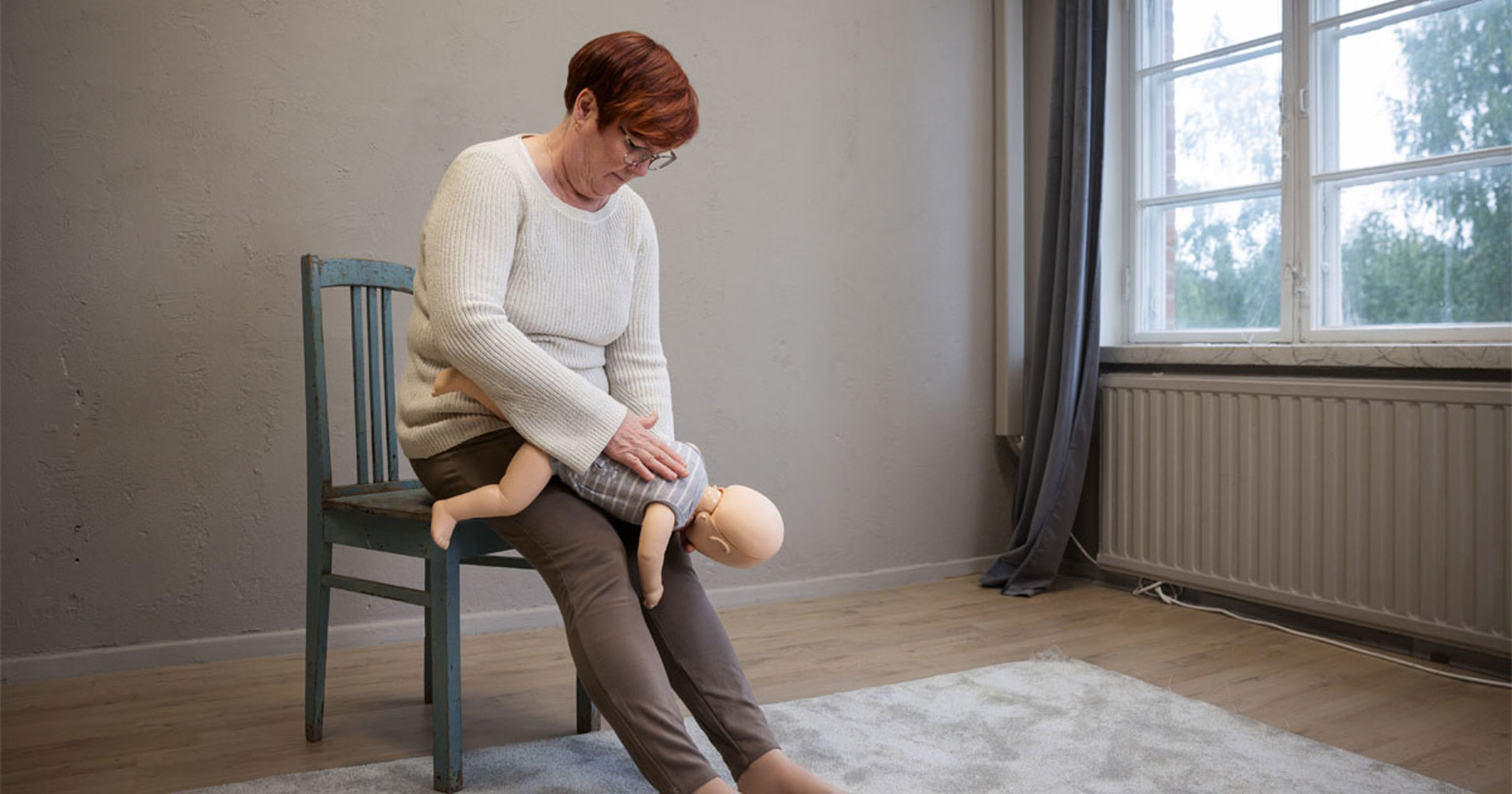 Henkilö istuu tuolilla pitäen käsissään vauva-elvytysnukkea ja iskee tätä selkään harjoitellen vierasesineen poistamista hengitysteistä.