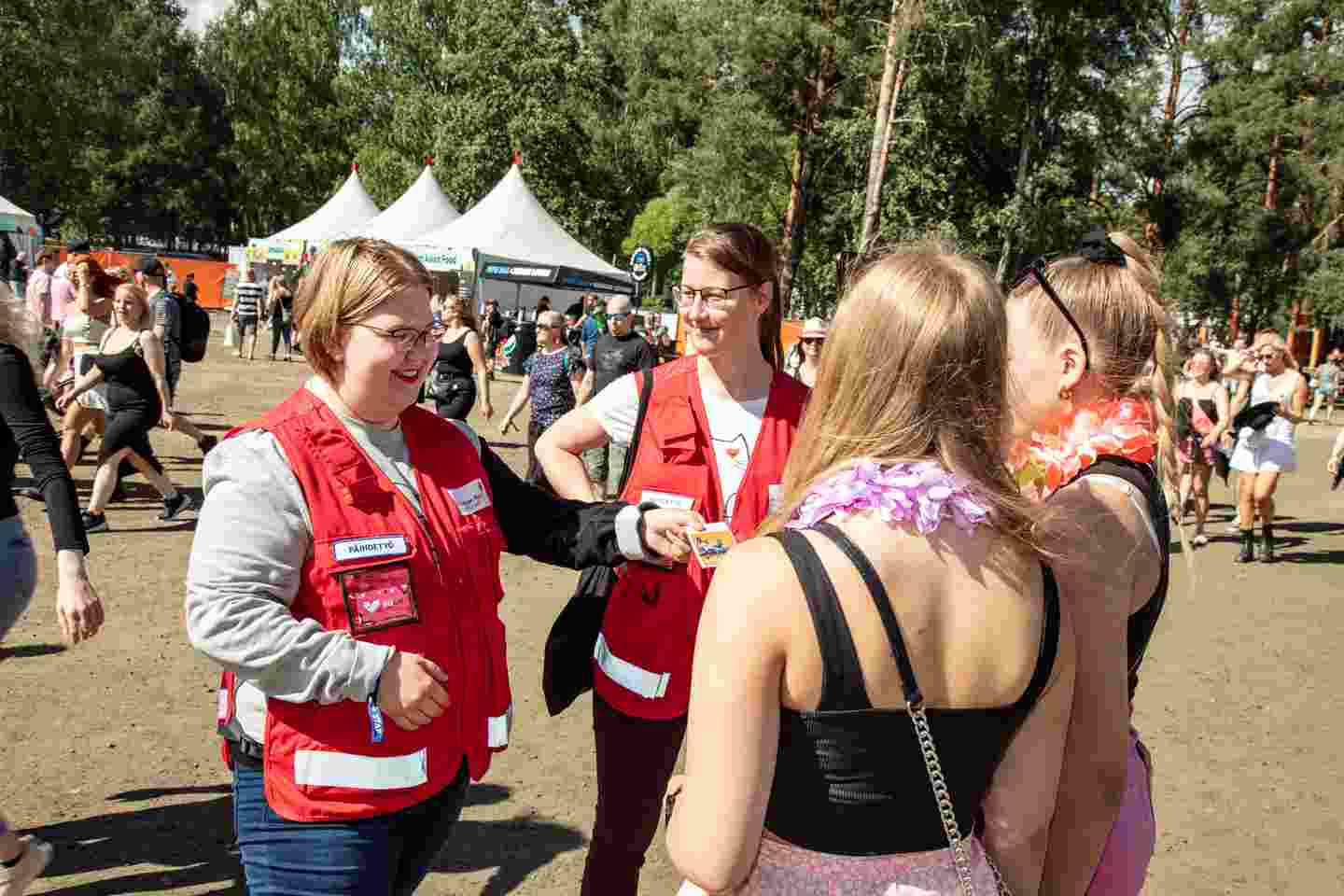 Punaisen Ristin vapaaehtoiset ojentavat kahdelle henkilölle esitteen. Taustalla aurinkoinen festarimiljöö.