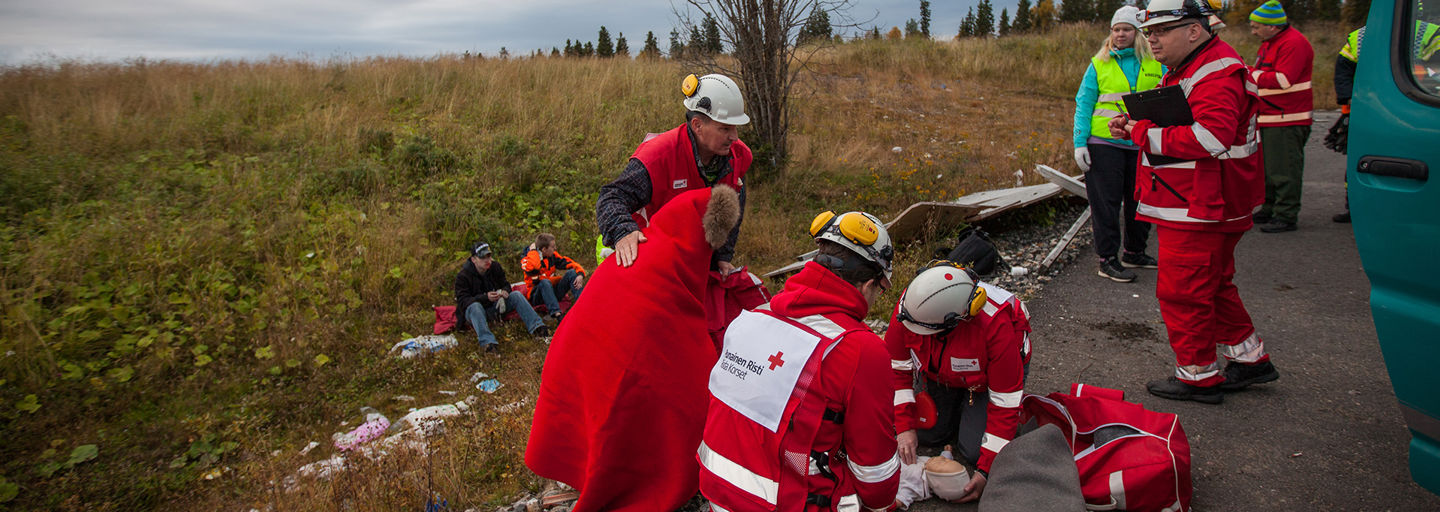Kaksi Punaisen Ristin rinnepäivystäjää käärii huopaa lumella makaavan henkilön ympärille laskettelurinteen reunassa.